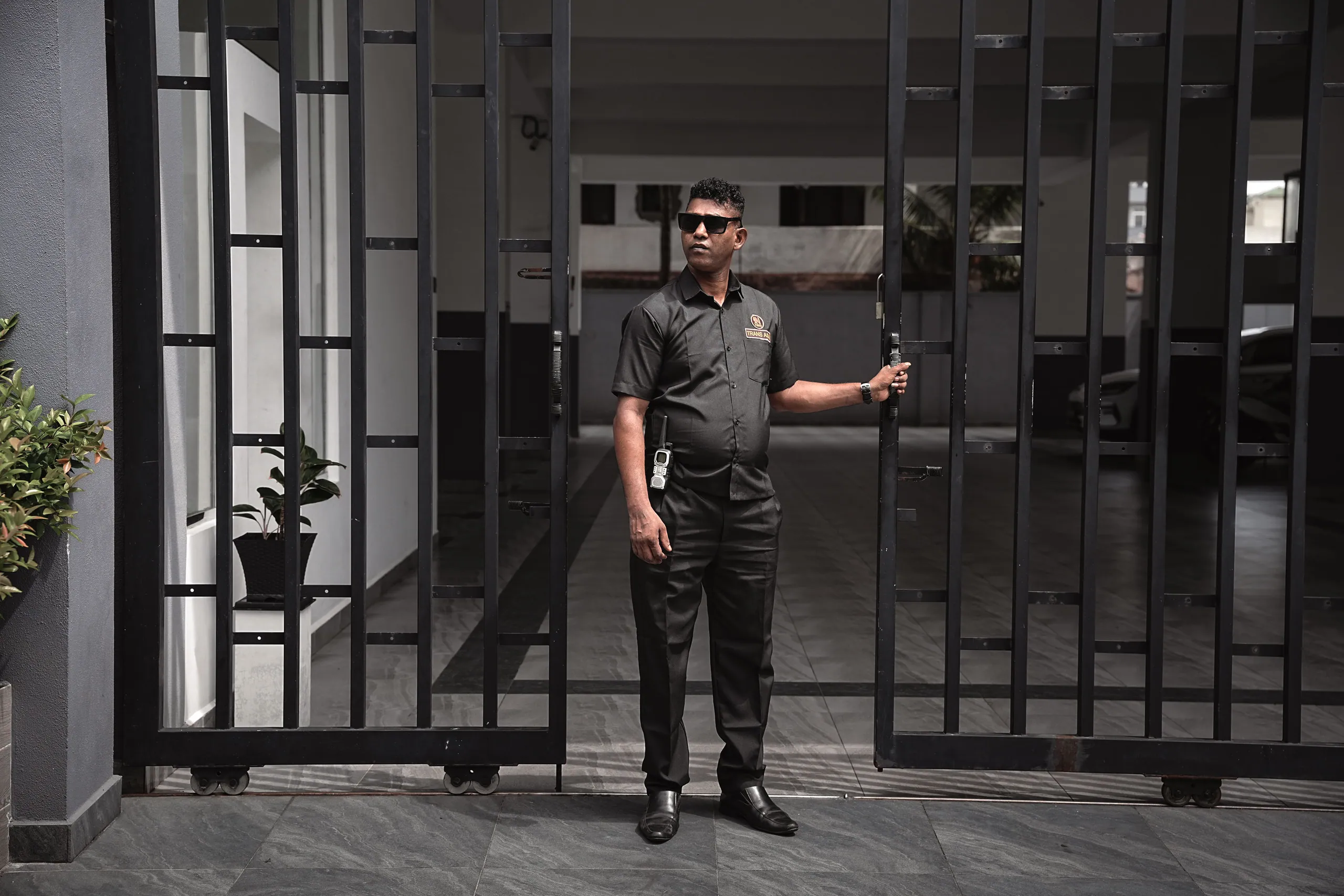 Trans Asia officer in black uniform standing in the open gate of a residence, one hand on the gate
