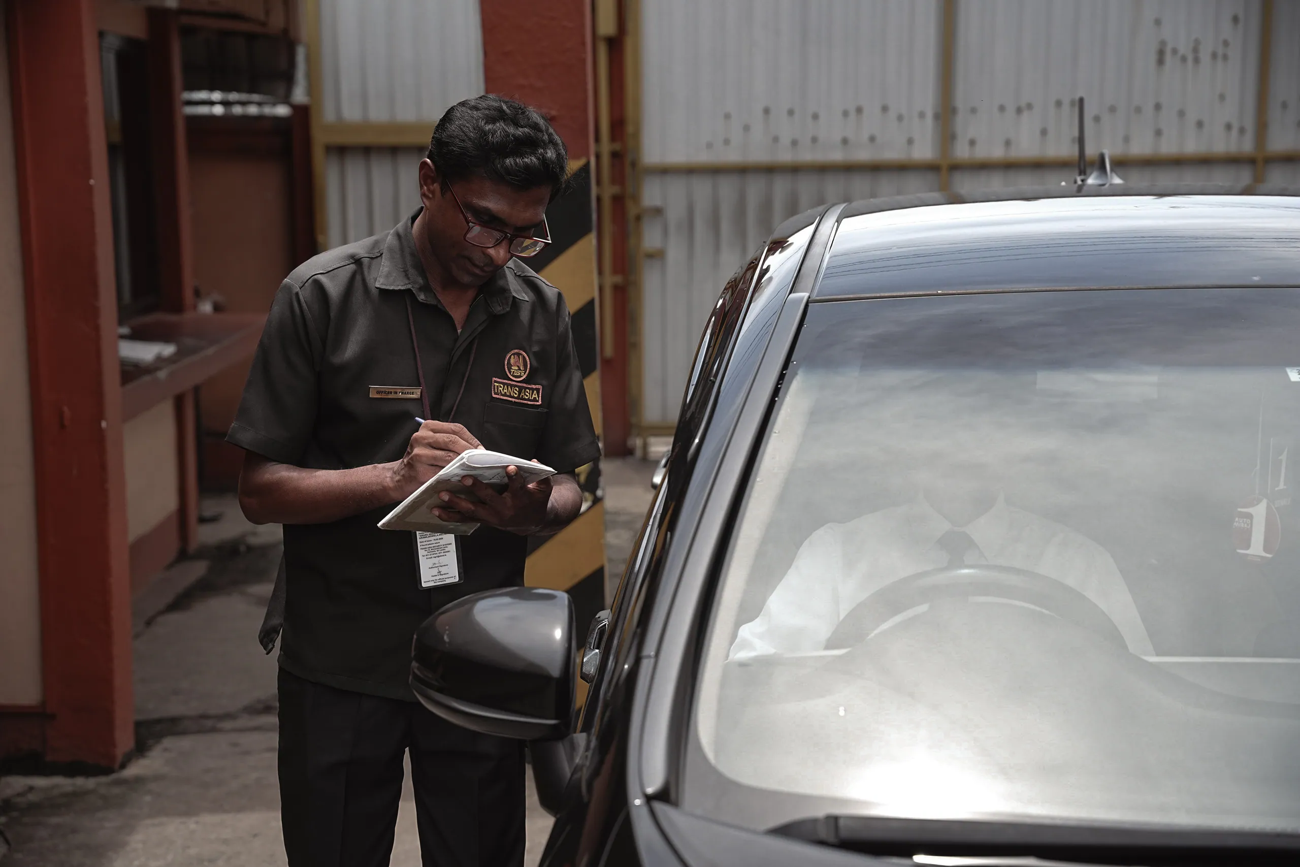 Officer in charge writing in a log book beside a car at a factory gate